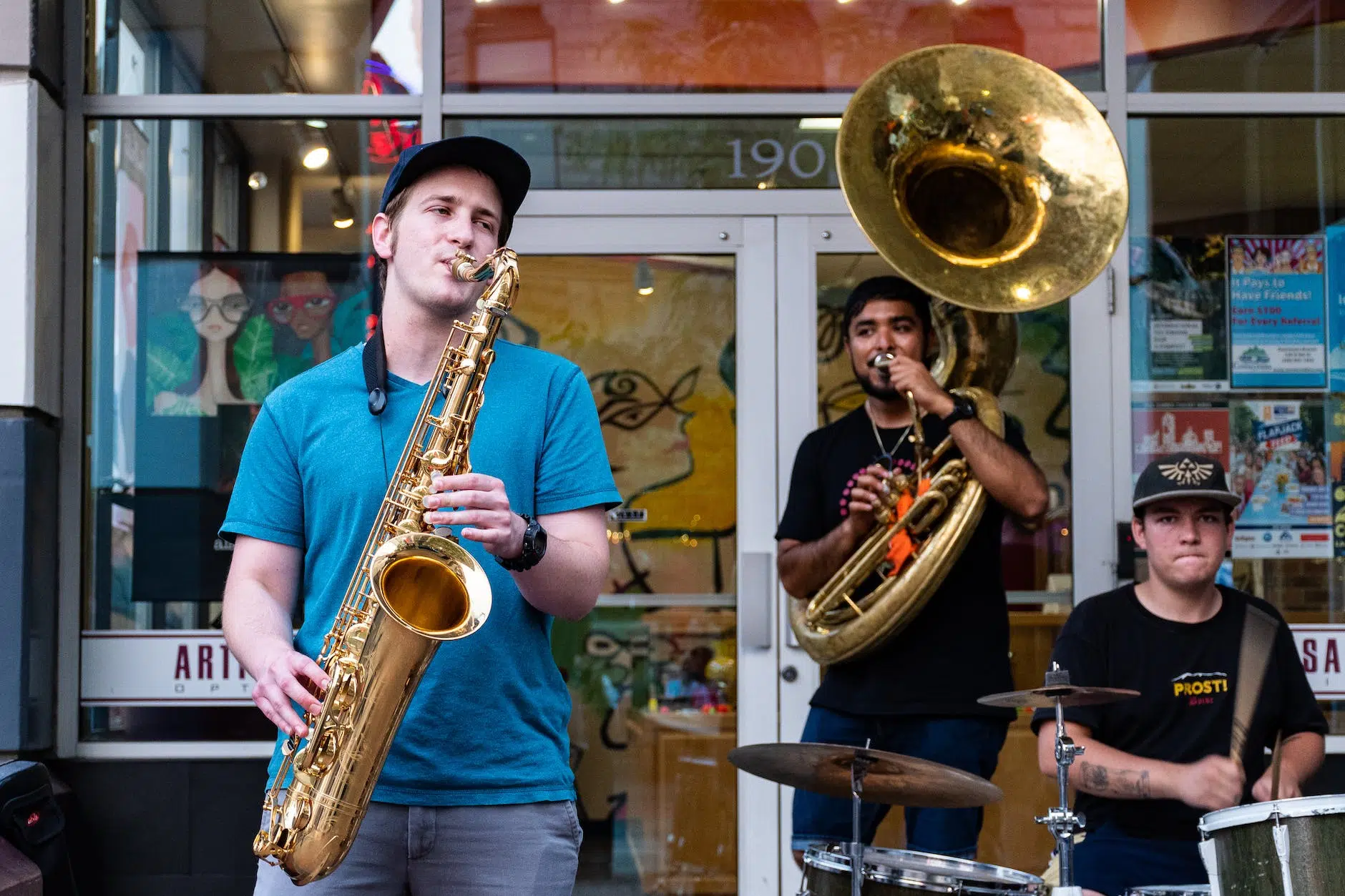 tres hombres tocando musica, saxo, trombón y batería en frente de una academia de arte